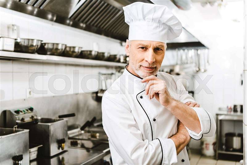 Handsome male chef in uniform and cap touching chin andf looking at camera in restaurant kitchen, stock photo