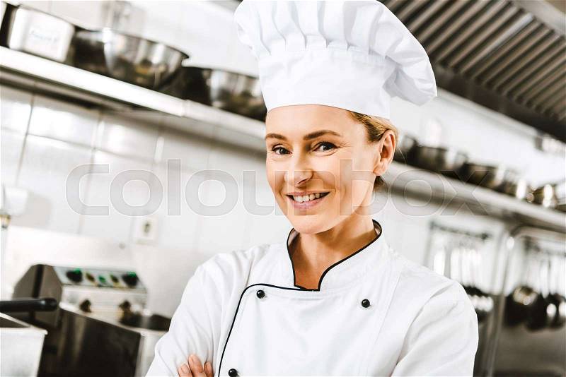 Beautiful smiling female chef in cap and double-breasted jacket looking at camera in restaurant kitchen, stock photo