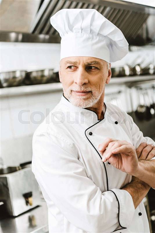 Handsome male chef in uniform and cap looking away in restaurant kitchen, stock photo