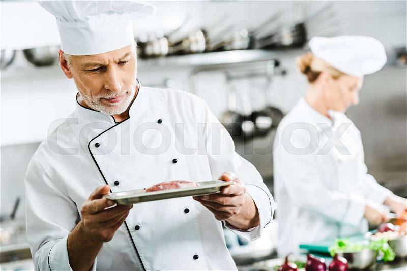 Pensive male chef in uniform holding meat dish on plate with colleague on background in restaurant kitchen, stock photo