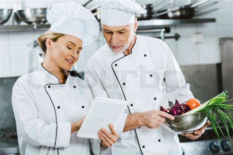 Female and male chefs in iniforms using digital tablet during cooking in restaurant kitchen, stock photo
