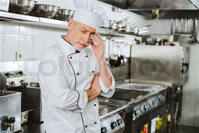 Tired male chef in uniform touching head and having headache in restaurant kitchen, stock photo