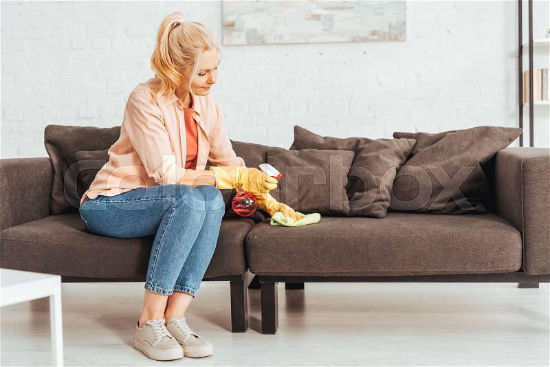 Senior woman in jeans cleaning sofa with spray and rag, stock photo
