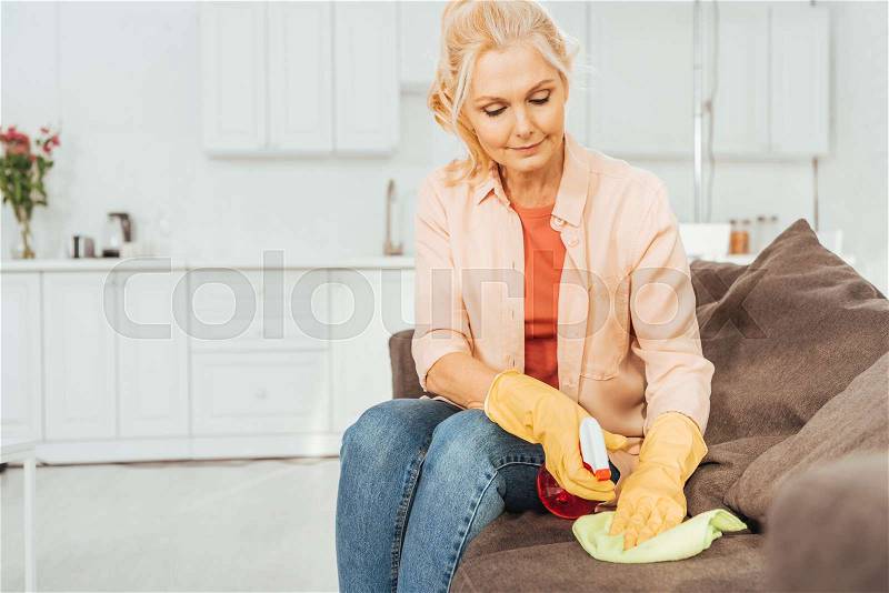 Senior woman in rubber gloves cleaning sofa with spray and rag, stock photo