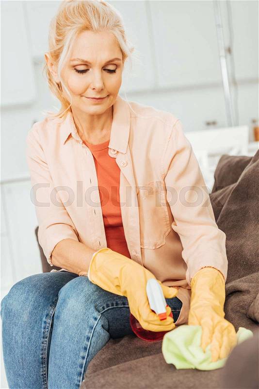 Senior woman in rubber gloves cleaning sofa with spray and rag, stock photo