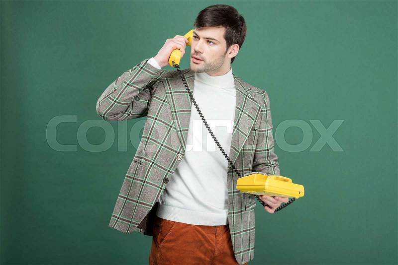 Handsome man in vintage clothes talking on retro rotary dial phone isolated on green, stock photo