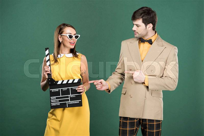 Man in vintage clothes pointing with fingers at woman with clapperboard isolated on green, stock photo