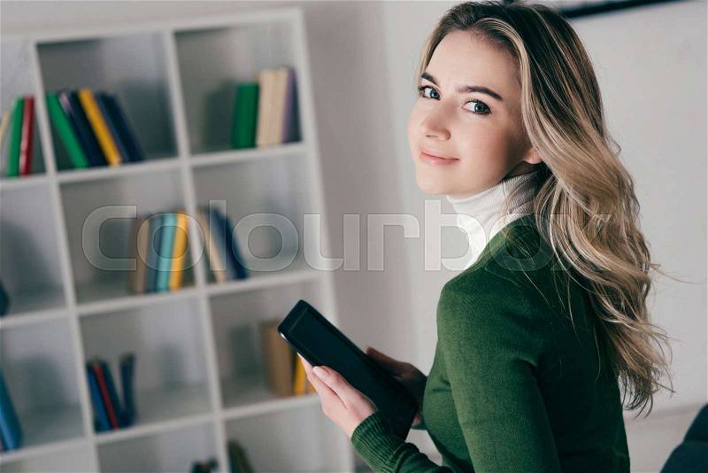 Cheerful woman holding e-reader and smiling near bookshelf, stock photo