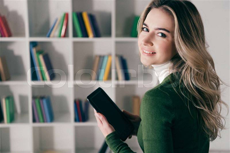 Happy woman holding e-book with blank screen near bookshelf at home , stock photo
