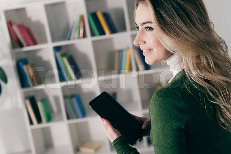Cheerful woman holding e-book with blank screen near bookshelf at home , stock photo