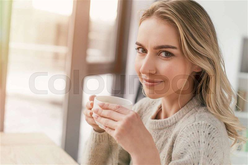 Happy woman holding cup with tea and looking at camera at home, stock photo