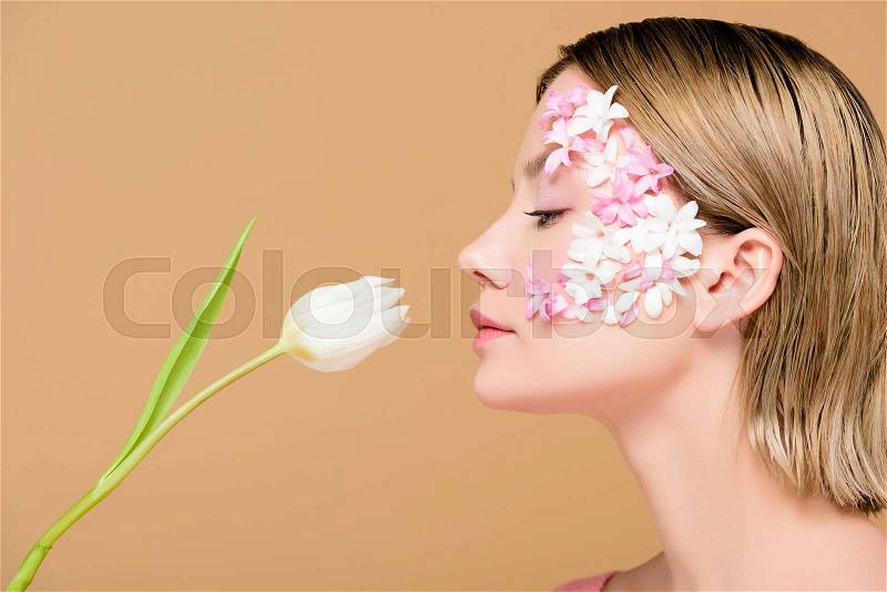 Side view of elegant woman with flowers on face smelling tulip isolated on beige, stock photo