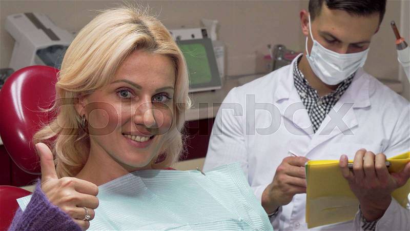 Happy female patient showing thumbs up sitting in dental chair at the dentists. Mature woman talking to her dentist while he is filling medical papers. Service, ..., stock photo