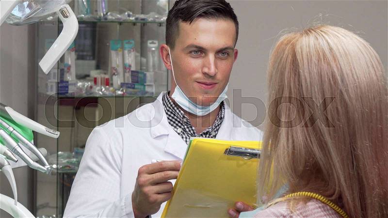 Young male dentist talking to his female patient, examining medical papers. Handsome male dentist smiling, checking papers of his patient, working at the dental ..., stock photo
