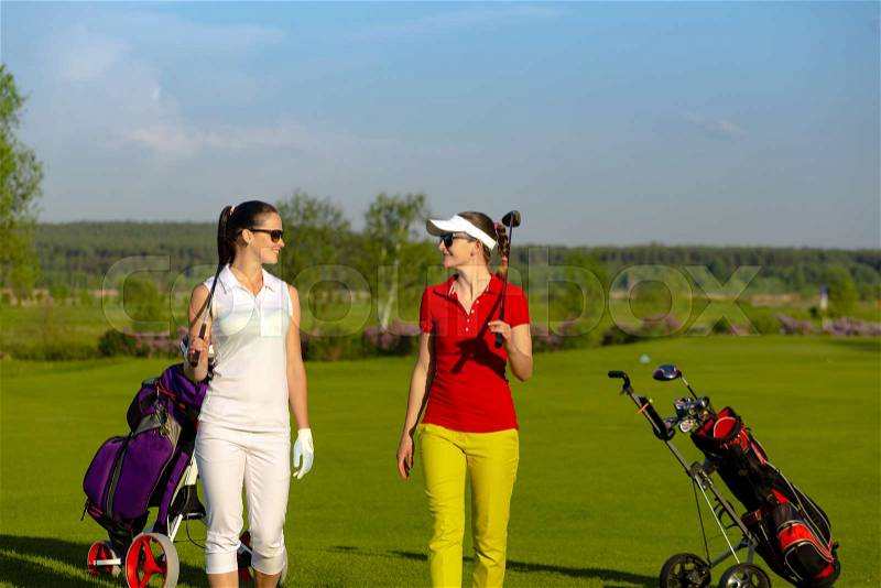 Two smiling athletic women golfers wearing sportwear and sunglasses walking on golf course at sunny day, stock photo