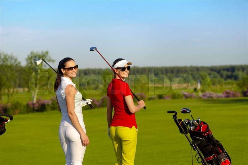 Two smiling athletic women golfers wearing sportwear and sunglasses walking and turn back on golf course at sunny day,, stock photo