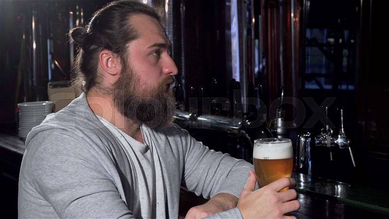 Bearded long haired man looking depressed, drinking beer alone at the bar. Hipster male sipping craft beer at the pub, looking disappointed. Stress, unhappy, ..., stock photo