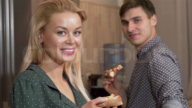 Stunning woman eating delicious pizza, smiling to the camera with her boyfriend. Lovely couple dining in their new apartment in the kitchen, smiling to the camera, ..., stock photo