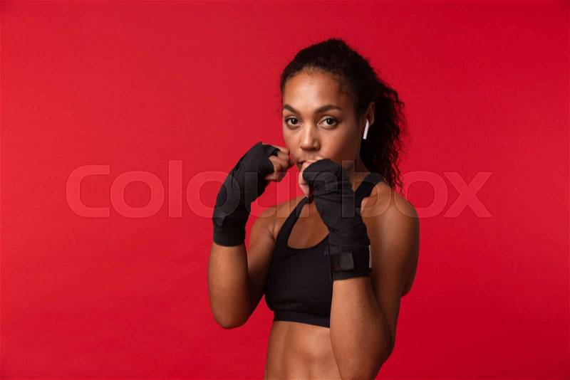 Portrait of strong african american woman in black sportswear boxing with sports bandages on her hands, isolated over red background, stock photo