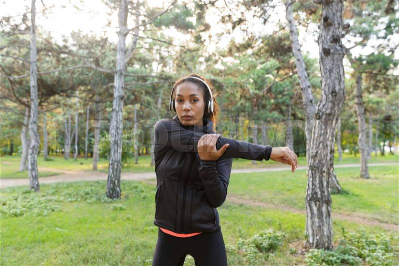 Image of athletic woman 20s wearing black tracksuit working out, and stretching body in green park, stock photo