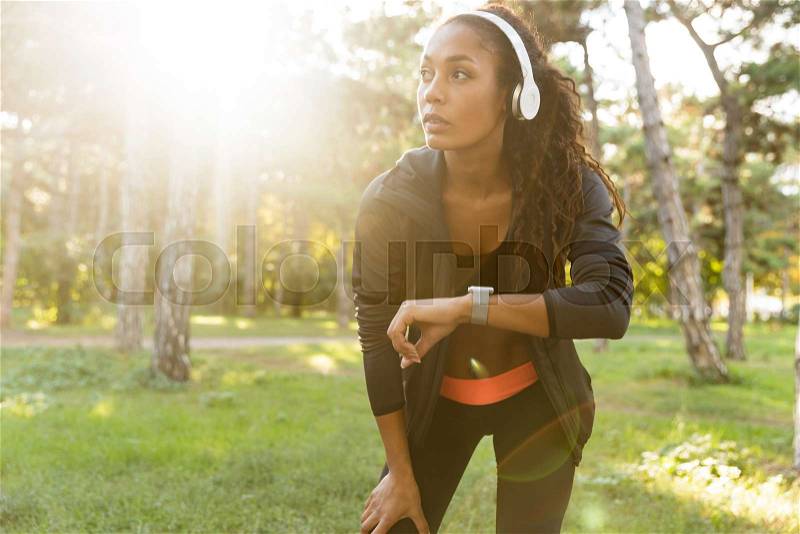 Image of skinny woman 20s wearing black tracksuit and headphones, walking through green park, stock photo