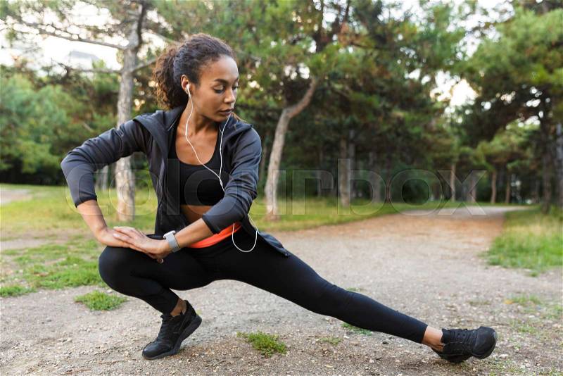 Portrait of well-built african american woman 20s wearing black tracksuit doing exercises, and stretching her legs in green park, stock photo
