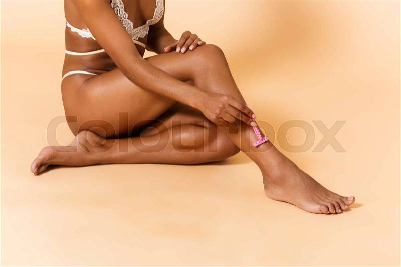 Beauty portrait of skinny woman wearing white lingerie shaving her legs, while sitting on the floor isolated over beige background, stock photo