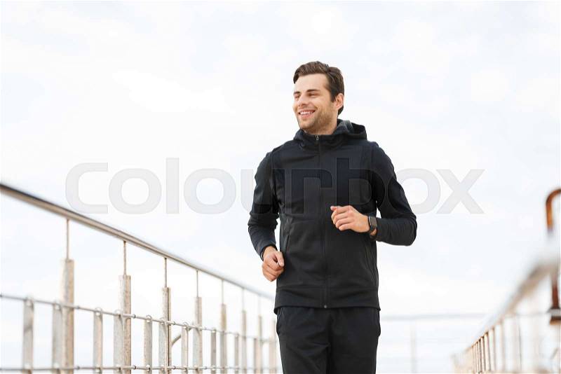 Image of joyful sporty man 30s in black sportswear, running along pier at seaside, stock photo