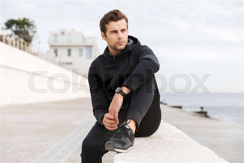 Image of handsome sporty man 30s in black sportswear, sitting on boardwalk at seaside, stock photo