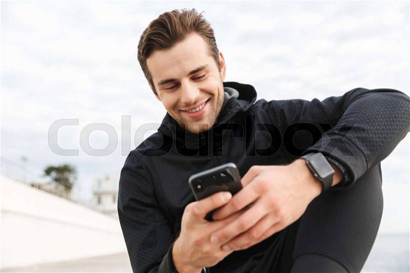 Image of sportive man 30s in black sportswear, using smartphone while sitting on boardwalk at seaside, stock photo