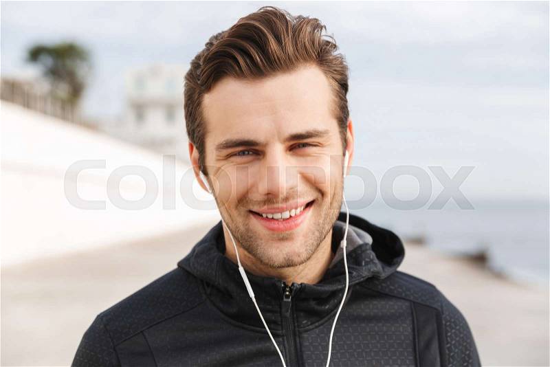 Image of sportive man 30s in black sportswear, using earphones and mobile phone while walking along boardwalk at seaside, stock photo