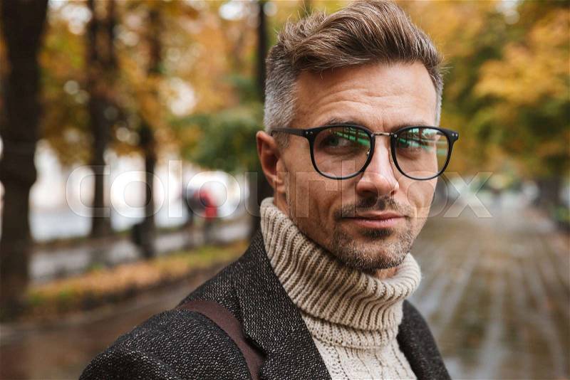 Photo of masculine man 30s wearing eyeglasses looking at camera, while walking outdoor through autumn park, stock photo