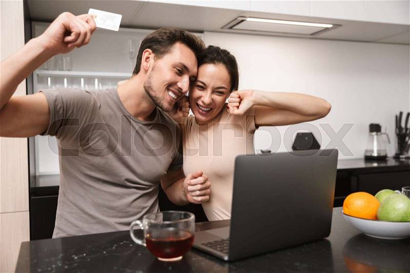 Photo of ecstatic couple man and woman using laptop with credit card, while sitting in kitchen, stock photo