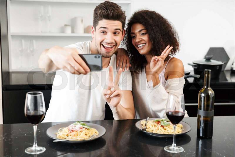 Beautiful young multiethnic couple having a romantic dinner at home, drinking red wine and eating pasta, toasting, taking a selfie, stock photo