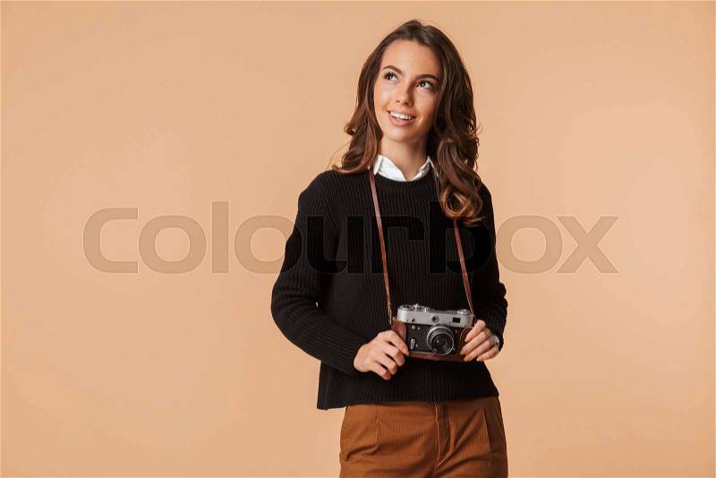 Pretty young woman wearing sweater holding photo camera isolated over beige background, looking away, stock photo