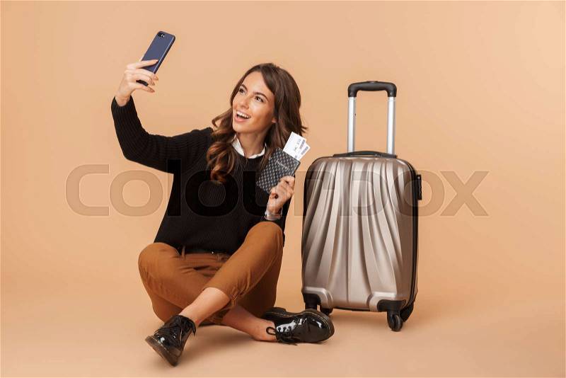 Young woman with baggage taking selfie photo on smartphone and holding travel ticket, while sitting on floor isolated over beige background, stock photo