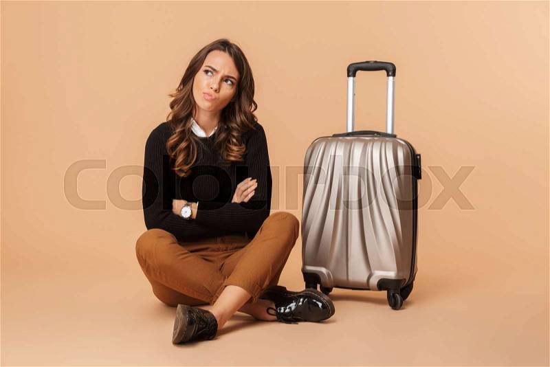 Photo of disappointed woman sitting on floor with baggage, while isolated over beige background, stock photo