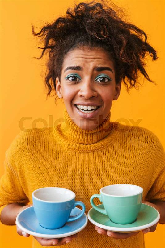 Confused african woman wearing sweater holding cups on saucers isolated over yellow background, stock photo