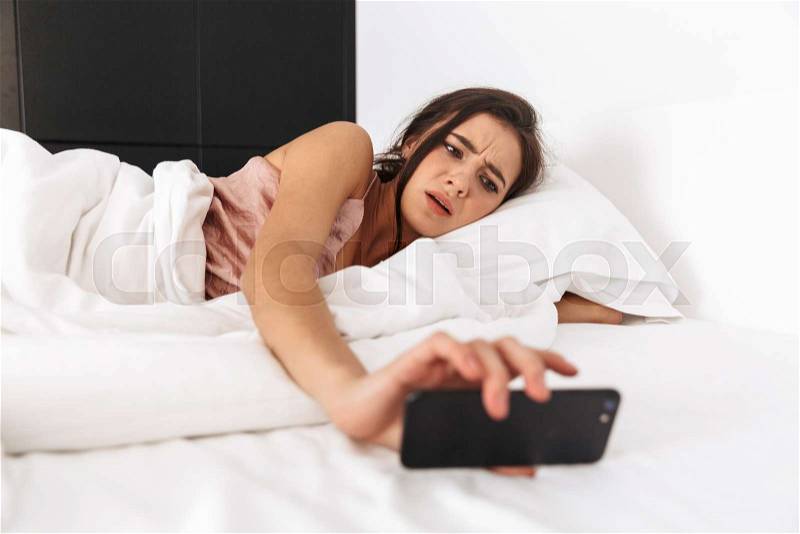Image of dissatisfied woman 20s looking at mobile phone, while sleeping in bed on white linen, stock photo