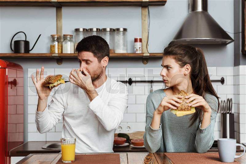 Disgusted young couple sitting at the kitchen during breakfast at home, holding sandwiches, stock photo