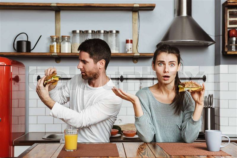 Disgusted young couple sitting at the kitchen during breakfast at home, holding sandwiches, stock photo