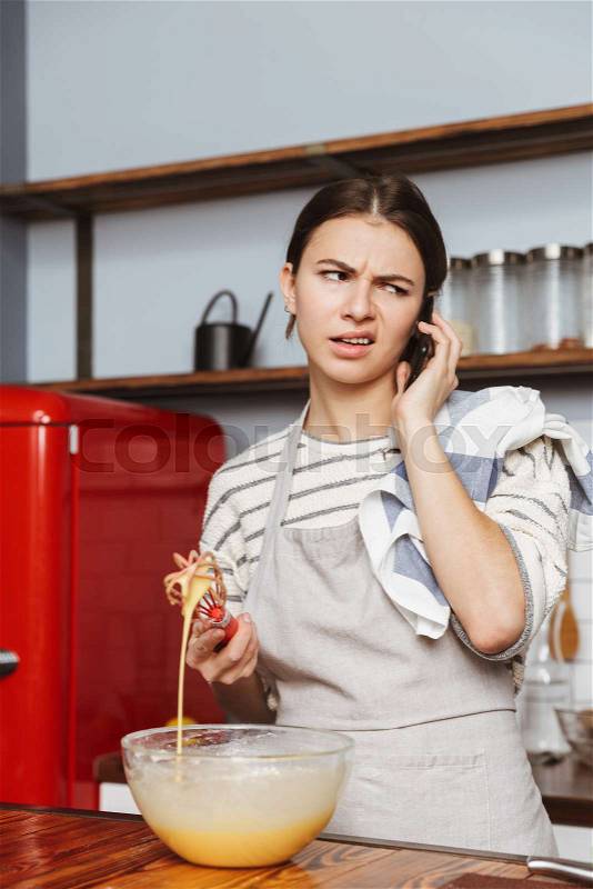 Confused young woman standing at the kitchen at home, mixing eggs in a bowl, talking on mobile phone, stock photo