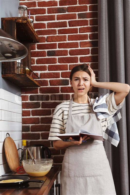Confused young woman standing at the kitchen at home, cooking pancakes on a frying pan, using mobile phone, stock photo