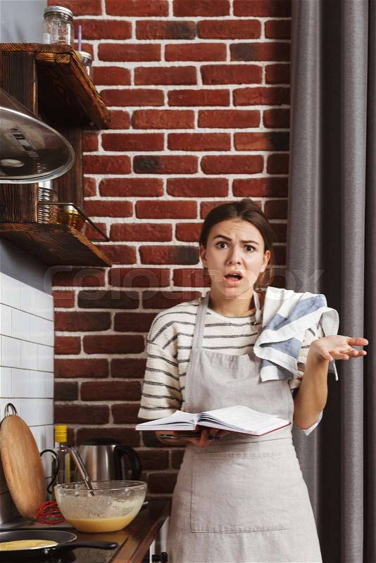 Confused young woman standing at the kitchen at home, cooking pancakes on a frying pan, using mobile phone, stock photo