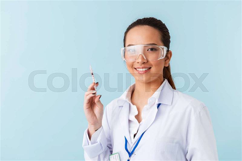 Photo of a young woman doctor posing isolated over blue wall background holding syringe, stock photo