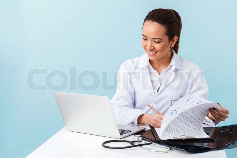 Photo of a young woman doctor posing isolated over blue wall background using laptop computer, stock photo