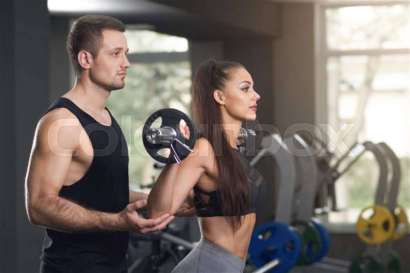 Professional personal trainer helping woman in gym with exercises. Beautiful brunette raising barbell, coach supporting. Charming woman with long hair wearing in ..., stock photo