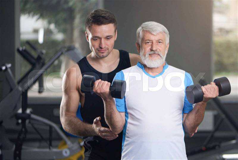 Elderly man with gray beard and hair posing in gym while working out. Old man holding dumbbells, looking at camera. Professional fit trainer helping pensioner with ..., stock photo