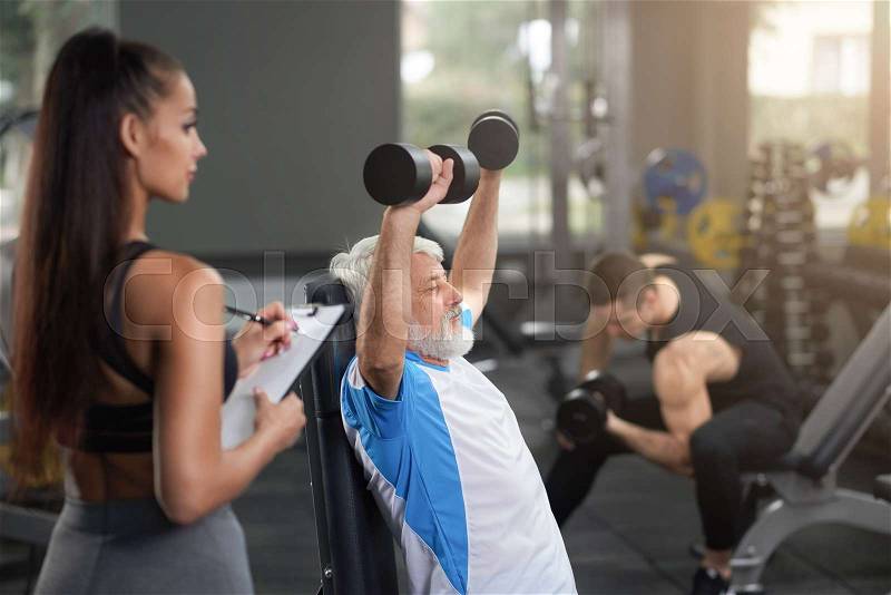 Healthy elderly man sitting, holding hands up and working out with dumbbells. Gorgeous brunette observing man, holding folder and writing with pen. Personal coach ..., stock photo