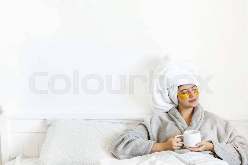 Beautiful young woman with eye patches in bathrobe holding cup of coffee and lying in bed, enjoying morning. Happy girl drinking coffee and applying eye patches, ..., stock photo